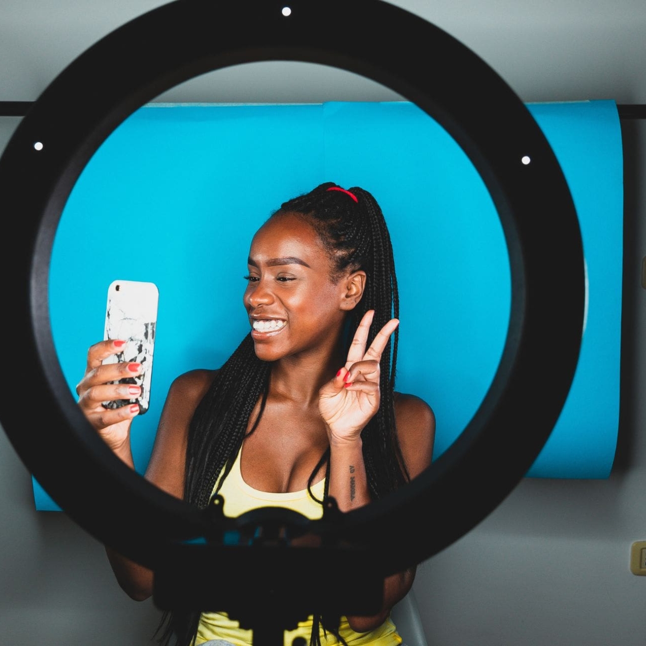 In a studio, a ring light frames a young Black female influencer who is smiling while looking into the mobile phone that she is holding.